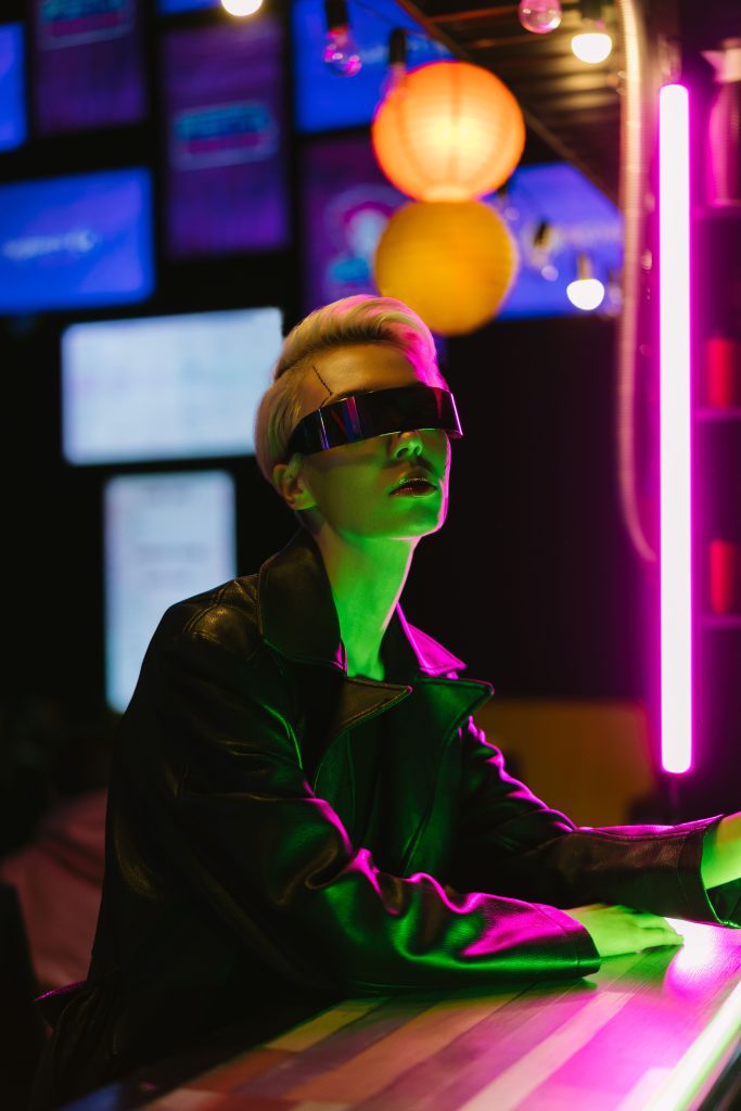Stylish woman in cyber sunglasses illuminated by neon lights, showcasing futuristic fashion.